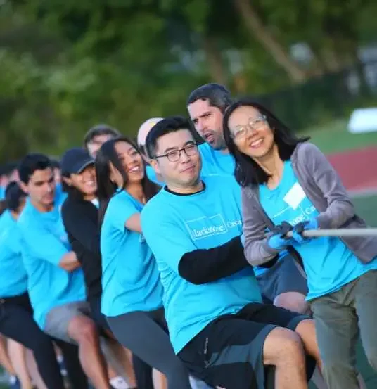 A group of people participating in a tug-of-war game outdoors, wearing matching blue shirts and smiling.