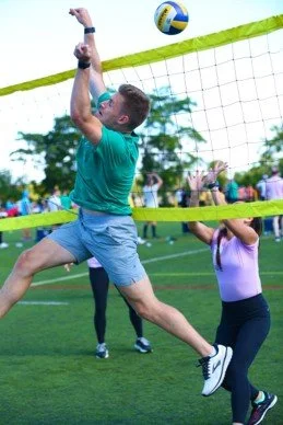 A man playing volleyball at a net outdoors with a woman, during daytime, with other people in the background.