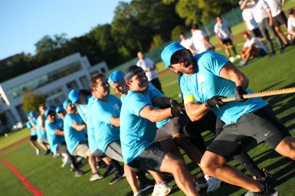 Group of people participating in a tug of war on a school track field with a building and spectators in the background