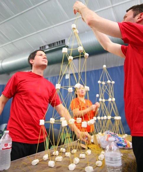 Three competitors wearing red and orange shirts building a structure with marshmallows and spaghetti sticks at a table, with another similar structure in the background.