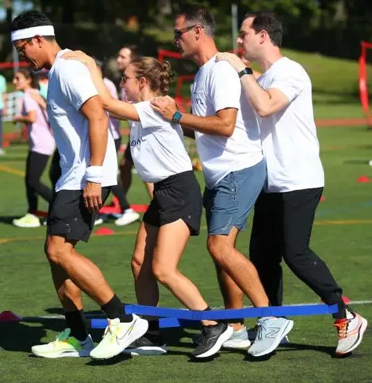 A group of four people participating in a fun team-building activity outdoors, walking in a line balancing on a blue slackline.