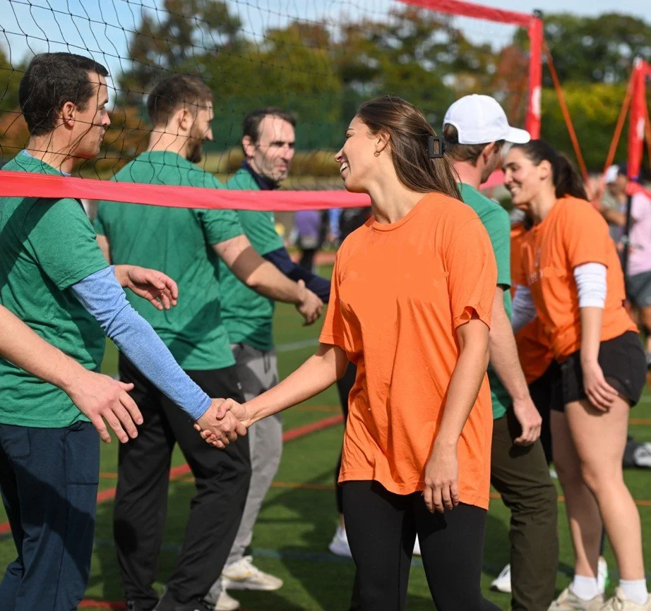 People shaking hands after a volleyball game on an outdoor court, some wearing orange shirts and others in green shirts.