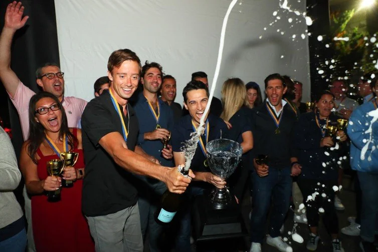 Group of people celebrating, some wearing medals, raising champagne bottles, and cheering with a large trophy on a table, with light-colored tent background.