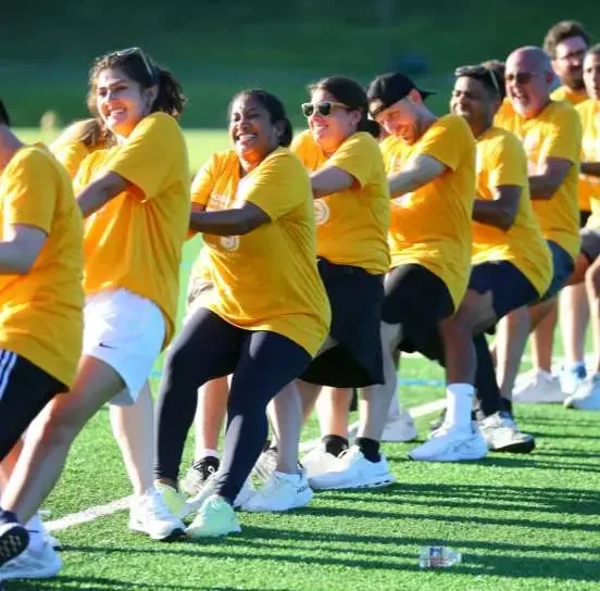 Group of diverse people in yellow shirts playing tug of war outdoors on a grassy field.