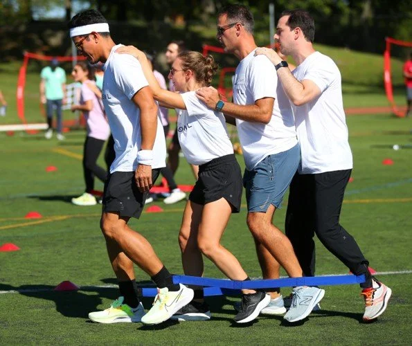 A group of four people participating in a team-building activity on a sports field, stepping over a blue rope held by two women in the background, with other participants and sports equipment visible.