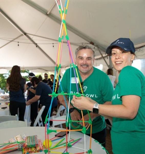Two people wearing green shirts under a tent, smiling, holding a colorful straw and pipe cleaner structure, with other people in the background