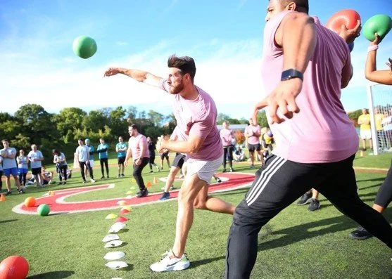 People participating in an outdoor athletic event, with a man throwing a green ball and others in the background, on a sports field with gym equipment and trees.