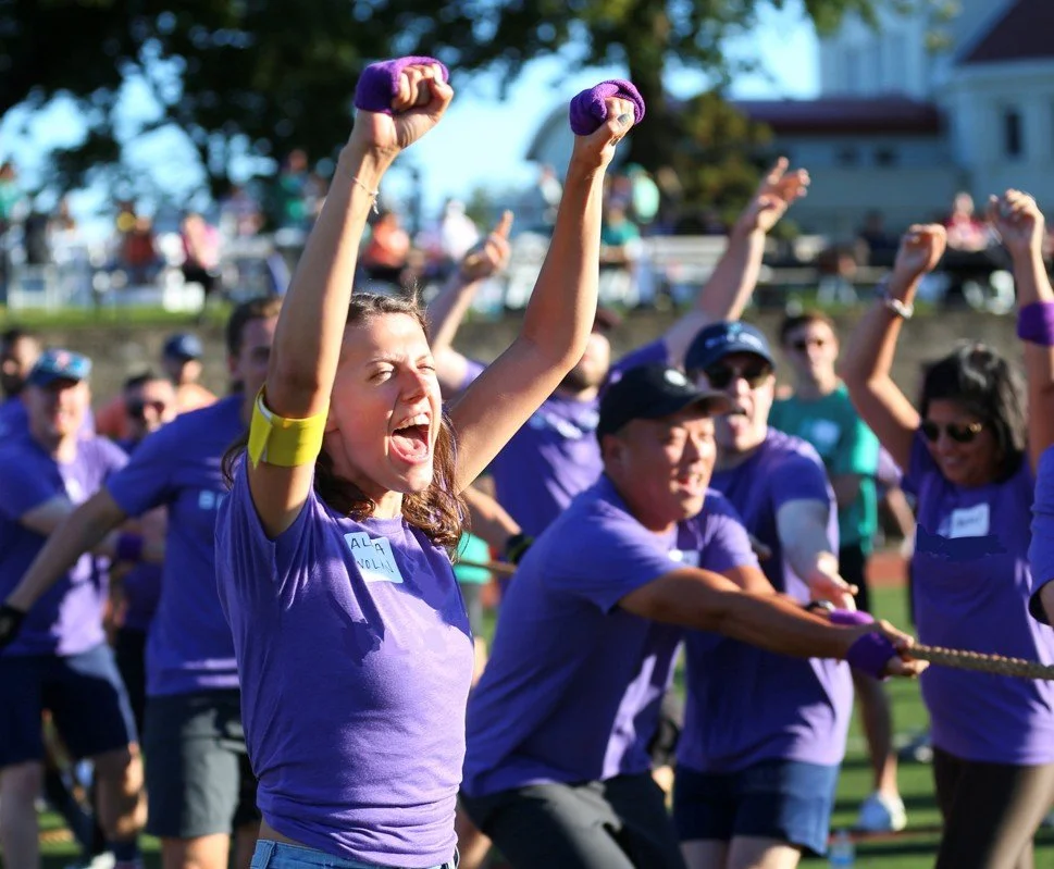 People participating in a tug-of-war contest outdoors, wearing purple shirts and raising their fists in victory.