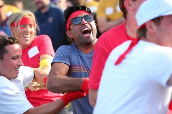People participating in a tug-of-war game, smiling and laughing, with some wearing red headbands and bands, outdoors during daytime.