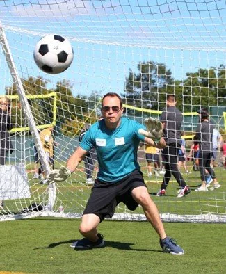 A man dressed as a goalkeeper in a teal shirt and black shorts is diving on a soccer field to catch a soccer ball.