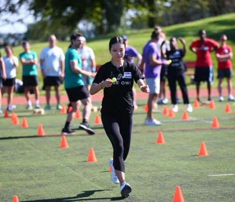 Girl running through orange cones set up for a drill on a grassy field with other people in the background.