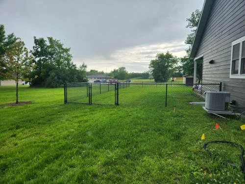 A grassy backyard with a black chain-link fence, an air conditioning unit, and small yellow and orange flags on the ground, under a cloudy sky.