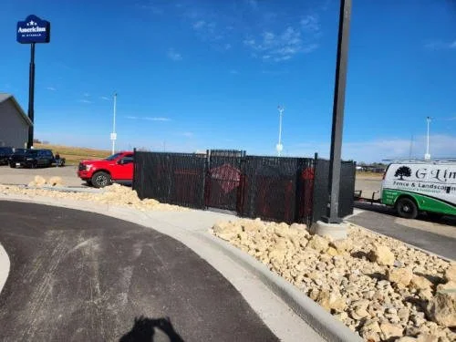 Parking lot with a red truck and a white utility vehicle, a black fence, rocks along the curb, and a blue sky.