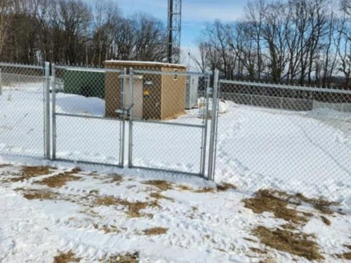 Chain-link fence with a double gate enclosing a small beige electrical or utility structure on snowy ground.