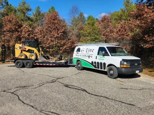 A landscaping company van attached to a trailer carrying a small yellow skid steer loader parked on a cracked asphalt surface with trees in fall colors in the background.