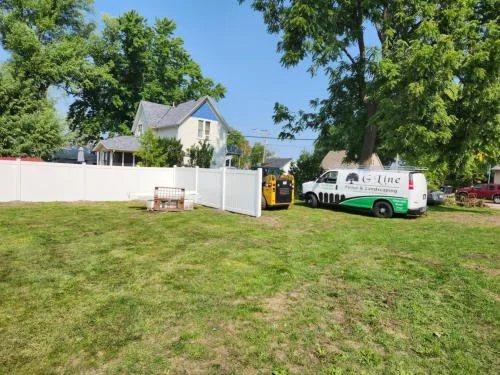 A grassy backyard with a white fence, a white van with green and black accents, a yellow generator, and a wooden bench, with trees and houses in the background.