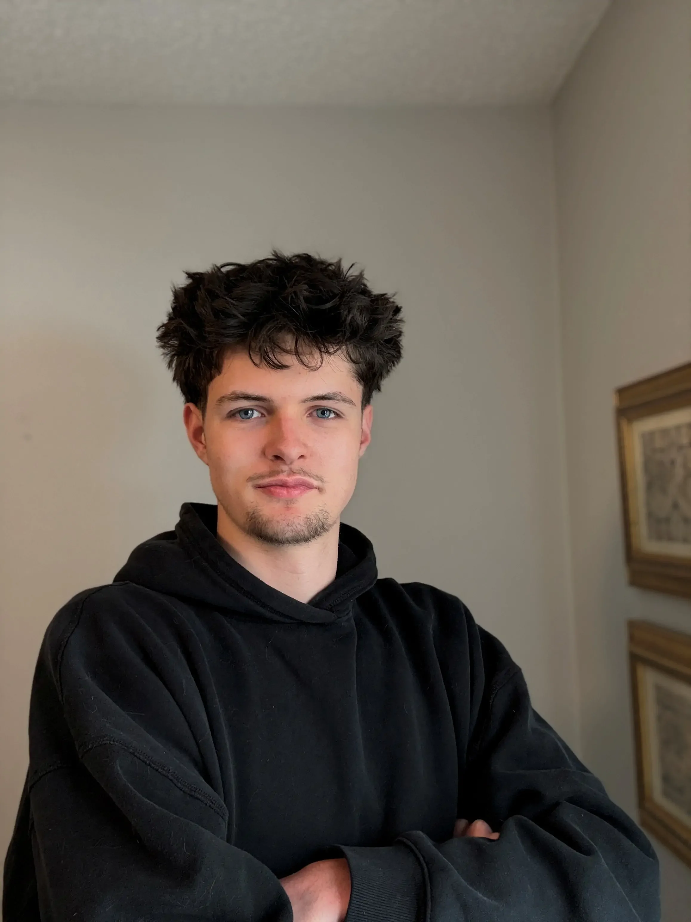A young man with dark, curly hair and a light beard, wearing a black hoodie, standing with arms crossed indoors against a plain wall with framed artwork