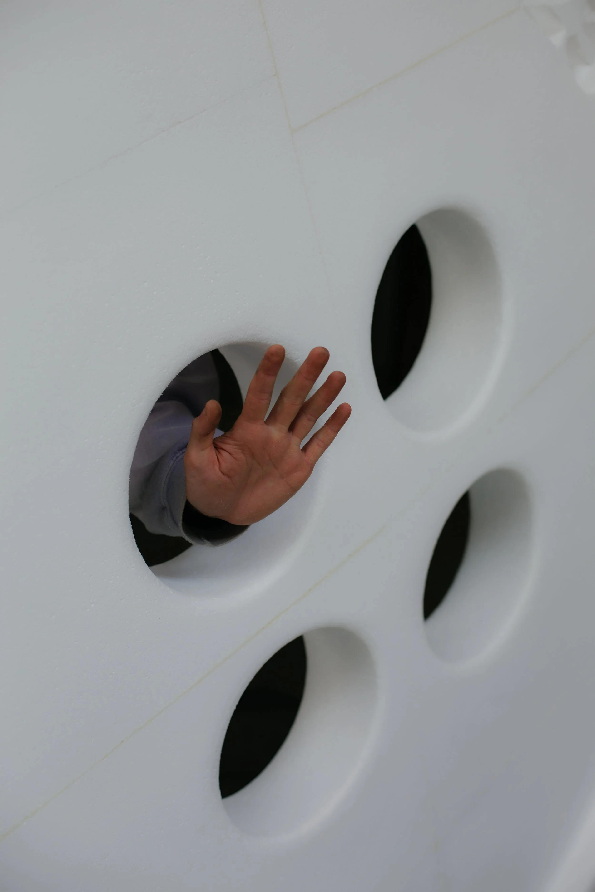Niños asomándose por círculos en una pared blanca con agujeros, jugando.