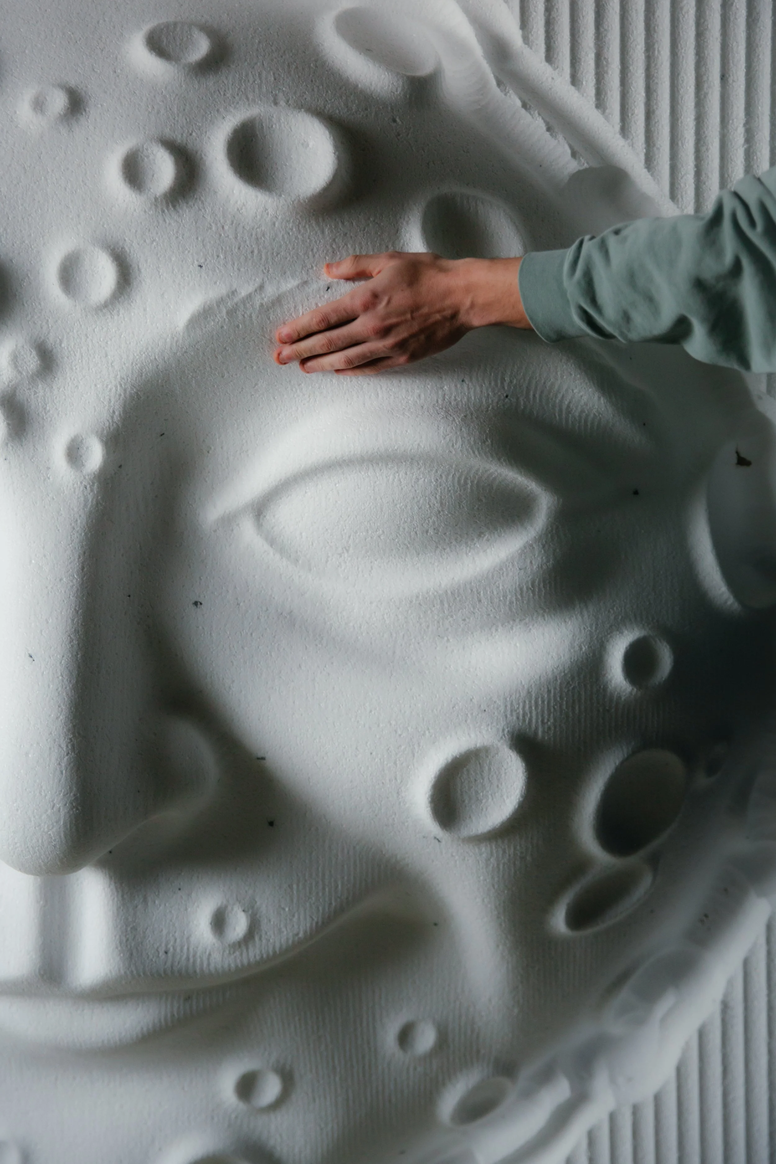 Persona tocando una escultura en relieve de una cara humana en una pared blanca.