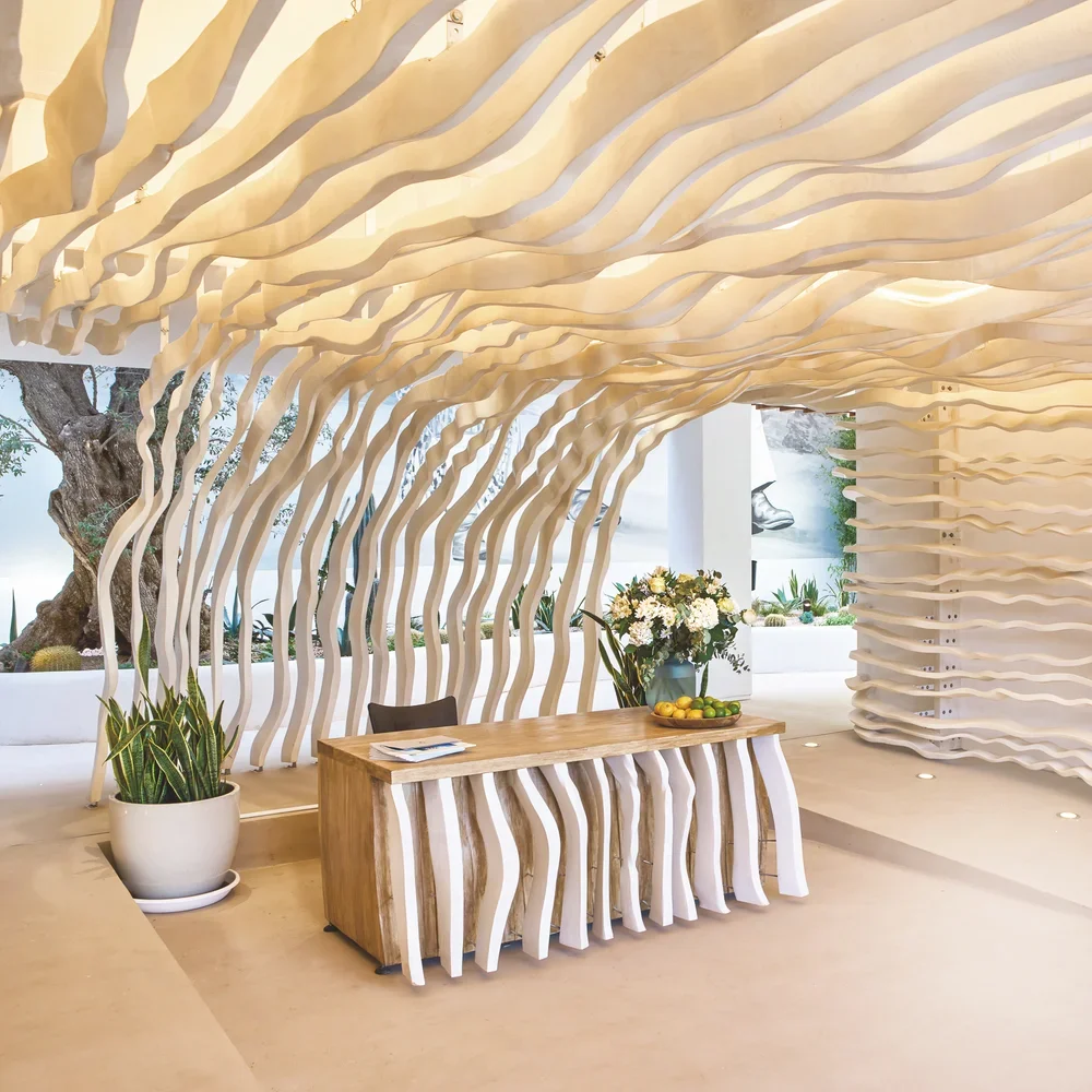 Estructura decorativa de madera con formas onduladas en un espacio interior, con mesa de madera, plantas y flores decorativas.