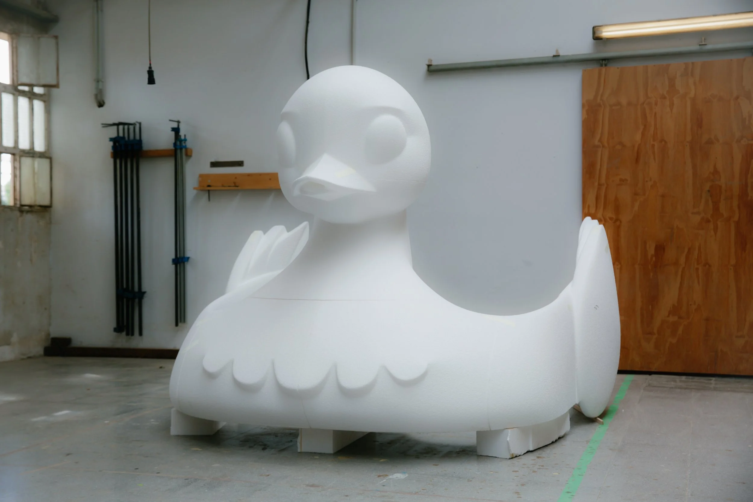 Escultura de una figura de un rostro grande y abstracto, de color blanco, ubicada en un espacio de taller o estudio con paredes blancas, una ventana, y accesorios de trabajo en el fondo.