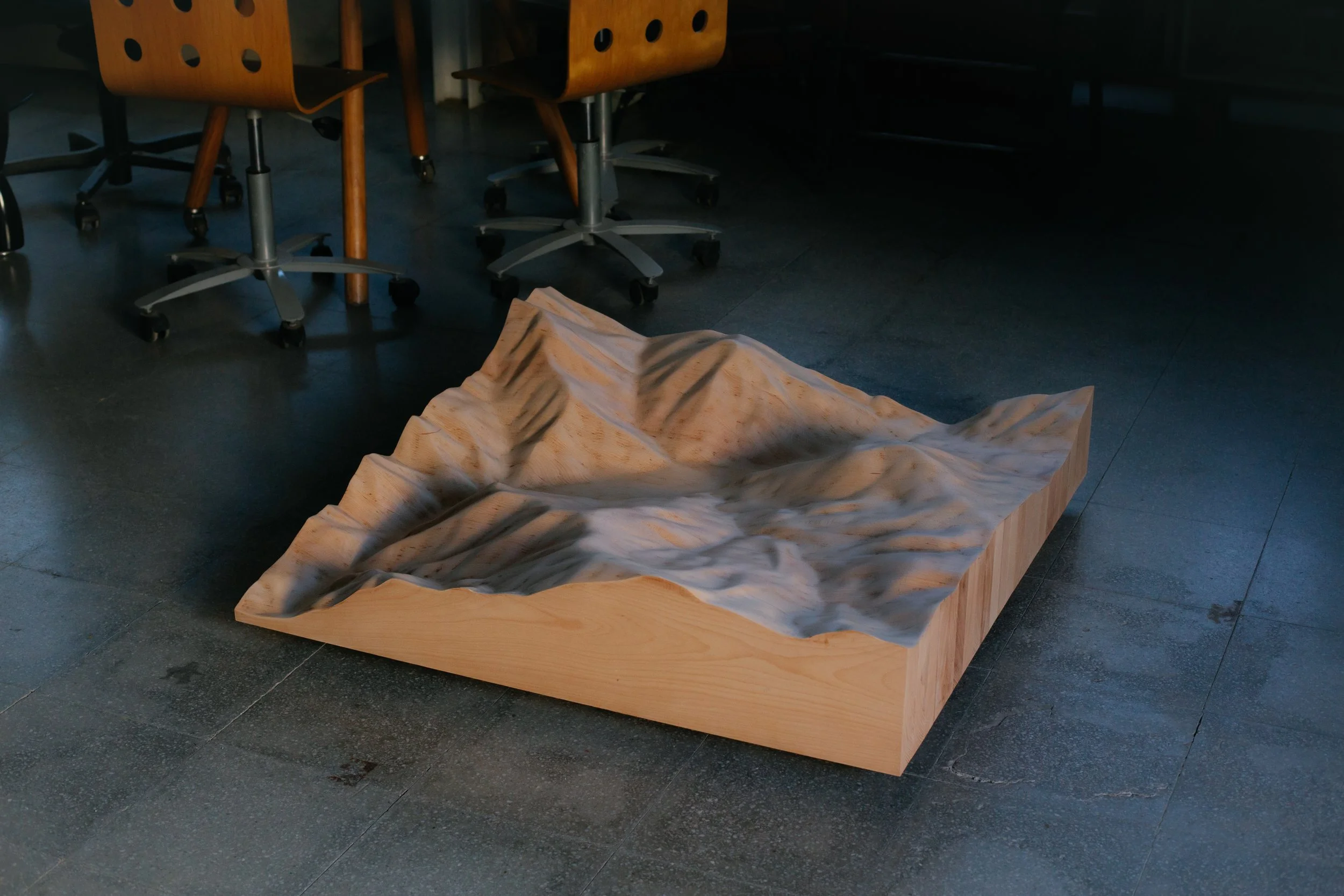 Escultura de madera que representa una montaña, ubicada en el suelo de un espacio interior con sillas de oficina y piso de baldosa oscura.