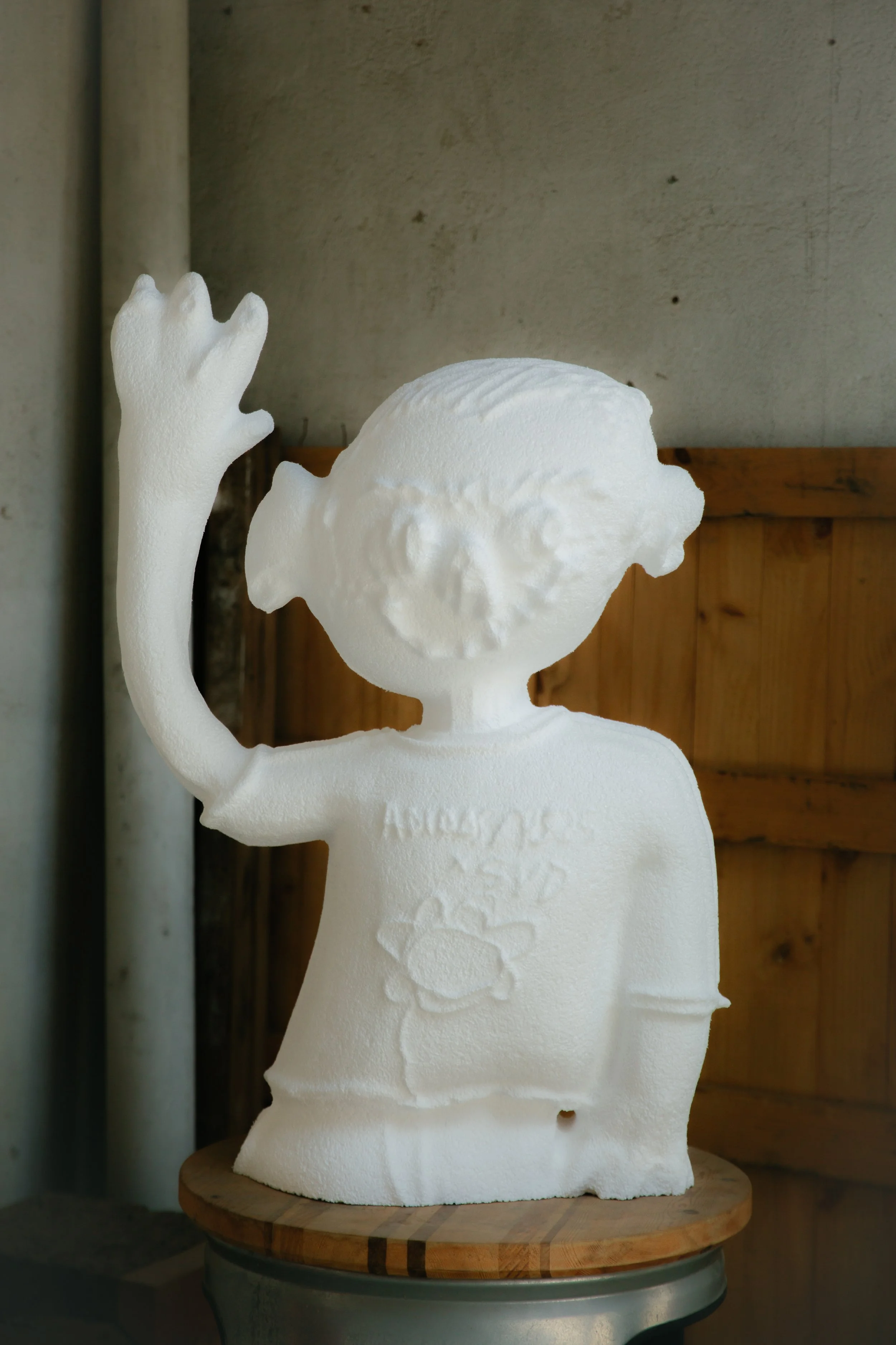 Escultura de una figura humana blanca con un brazo levantado, en forma de mano, y cabello rizado, en un fondo de pared de concreto y madera.