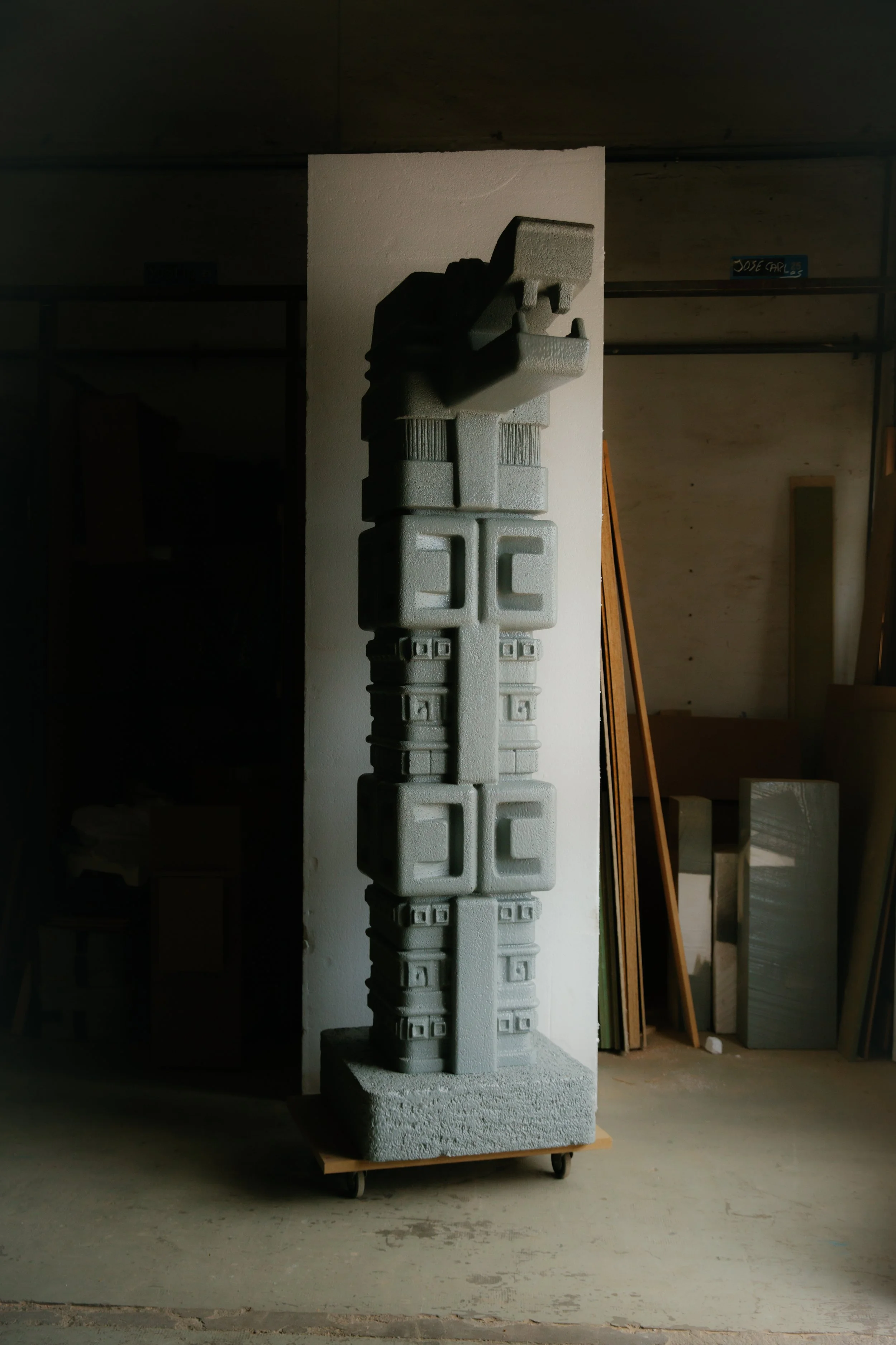 Escultura de piedra en forma de figura antropomorfa, ubicada en un espacio de construcción con paredes de madera y herramientas de carpintería en el fondo.