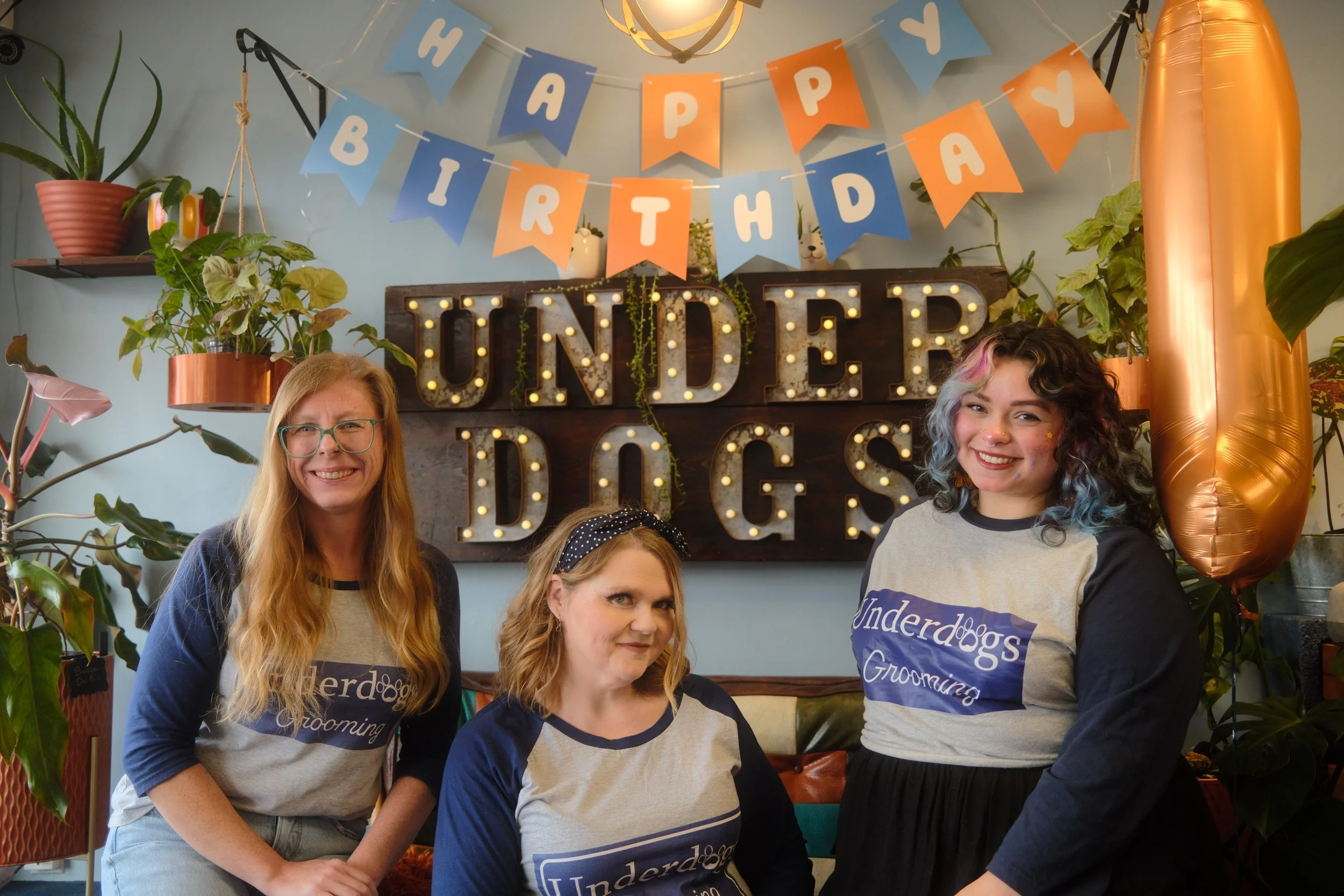 Three women celebrating a birthday in a cozy, decorated space with plants, a banner reading 'Happy Birthday,' and a marquee sign spelling 'Underdogs' with light bulbs. One woman has long blonde hair and glasses, another has shoulder-length hair with a polka-dot headband, and the third has curly hair with rainbow colors.