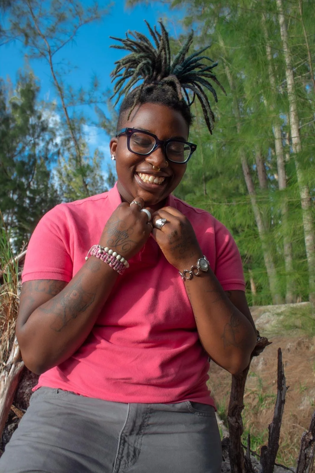 A woman smiling outdoors with trees and blue sky in the background. She has dreadlocks styled upwards, wears glasses, a pink shirt, and various rings and bracelets.