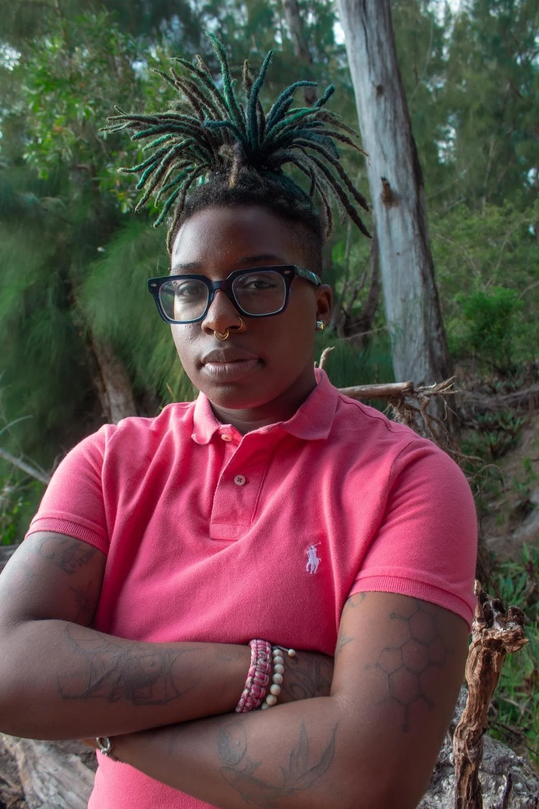 A person with tattoos on their arms and shoulder, wearing glasses, a septum piercing, and a pink polo shirt, stands outdoors with arms crossed in front of a forest background with trees and green foliage.
