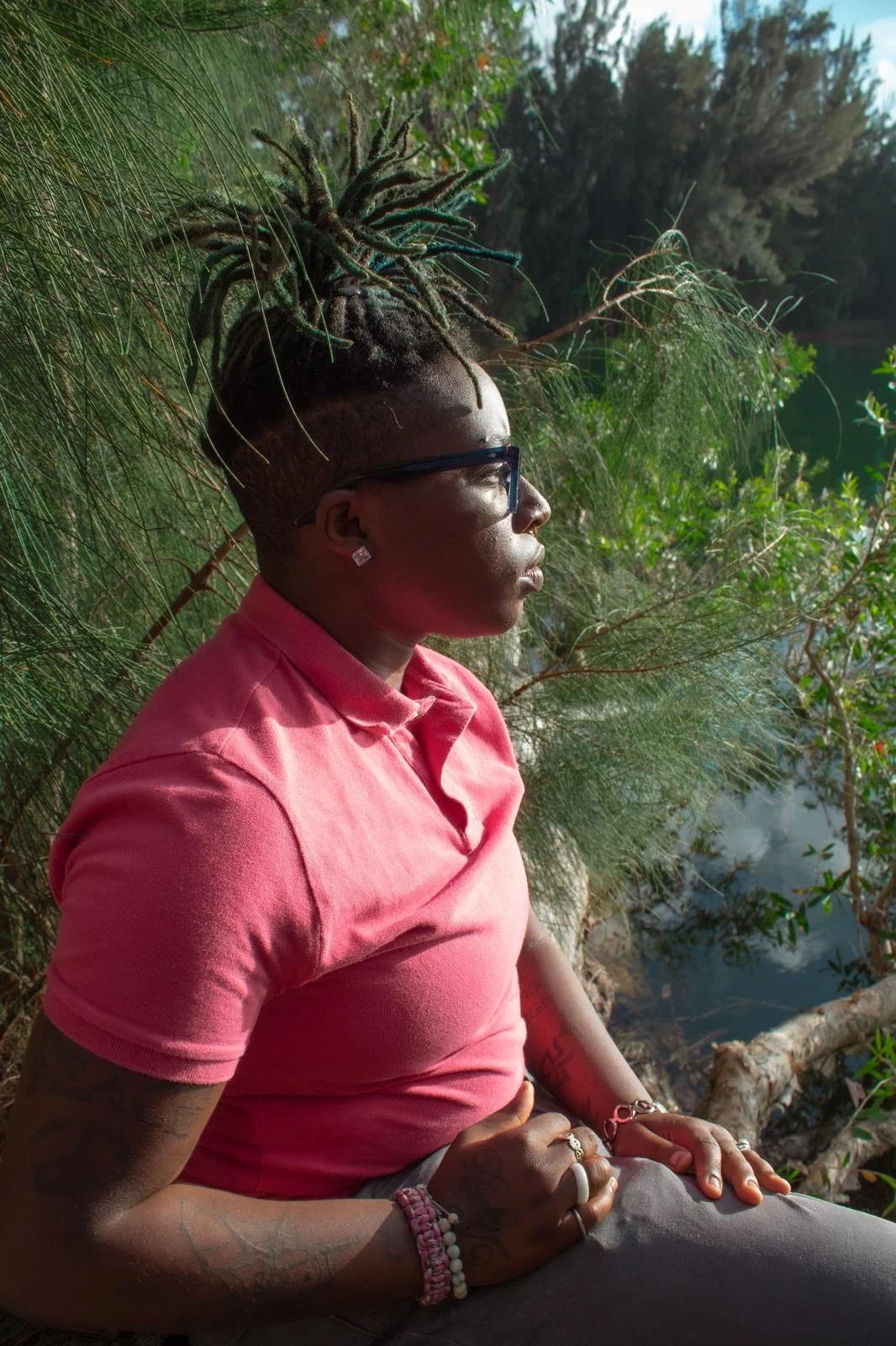 A woman with dreadlocks tied up on top of her head, wearing glasses, earrings, and a pink shirt, sitting outdoors near water surrounded by greenery.