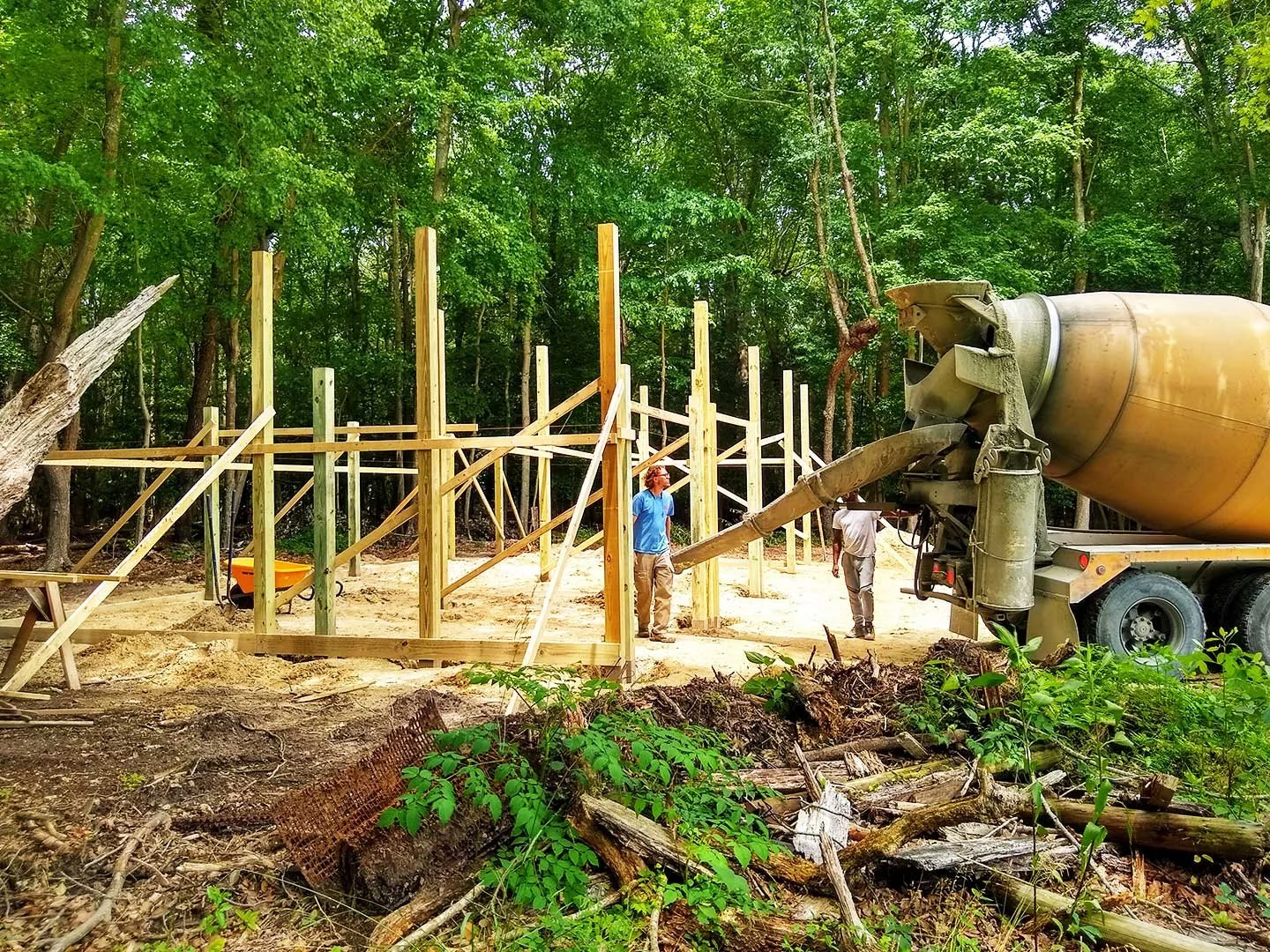 Construction site with partial wood framing in a wooded area where workers are pouring concrete from a cement truck.