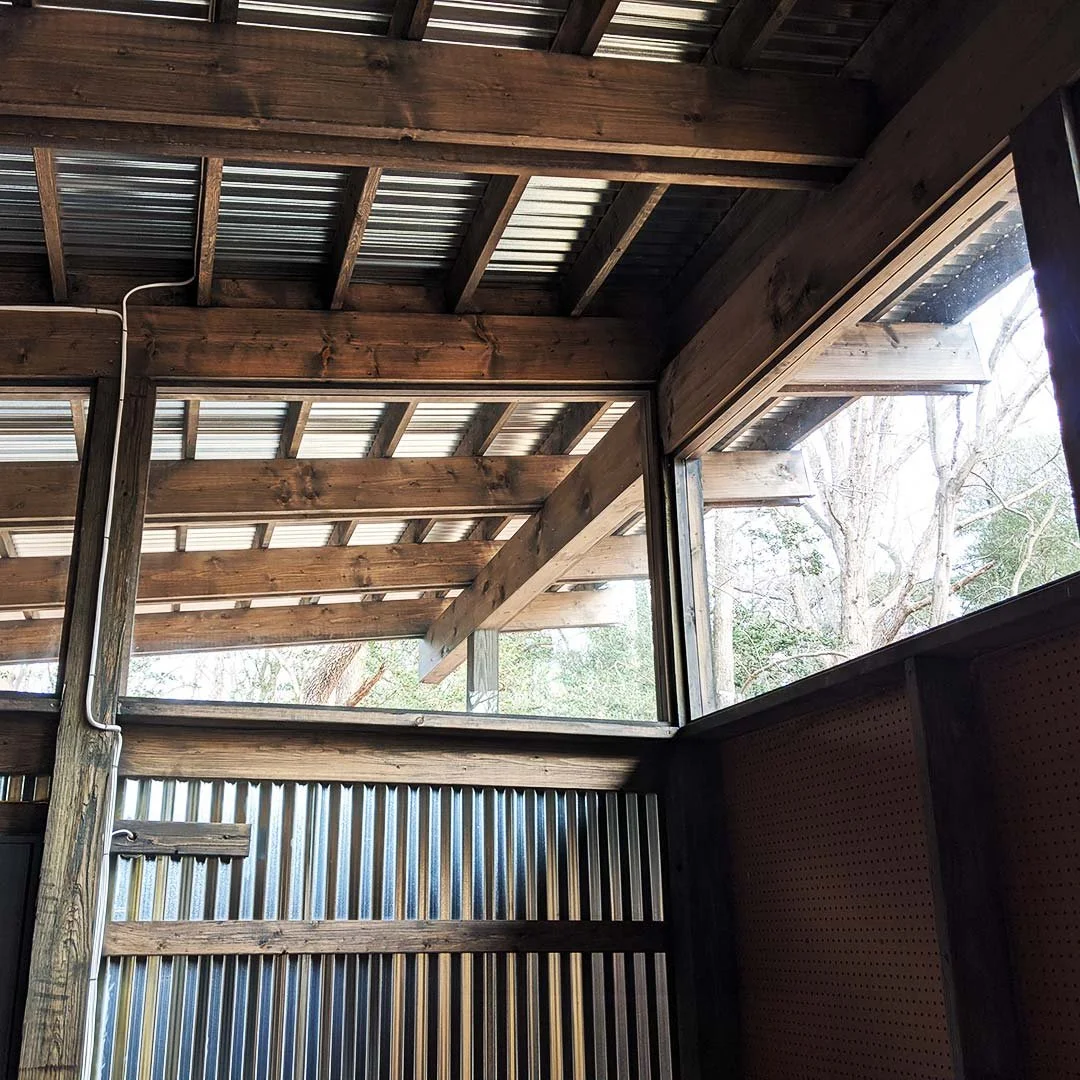 Interior view of a wooden and metal structure with large windows showing trees outside.