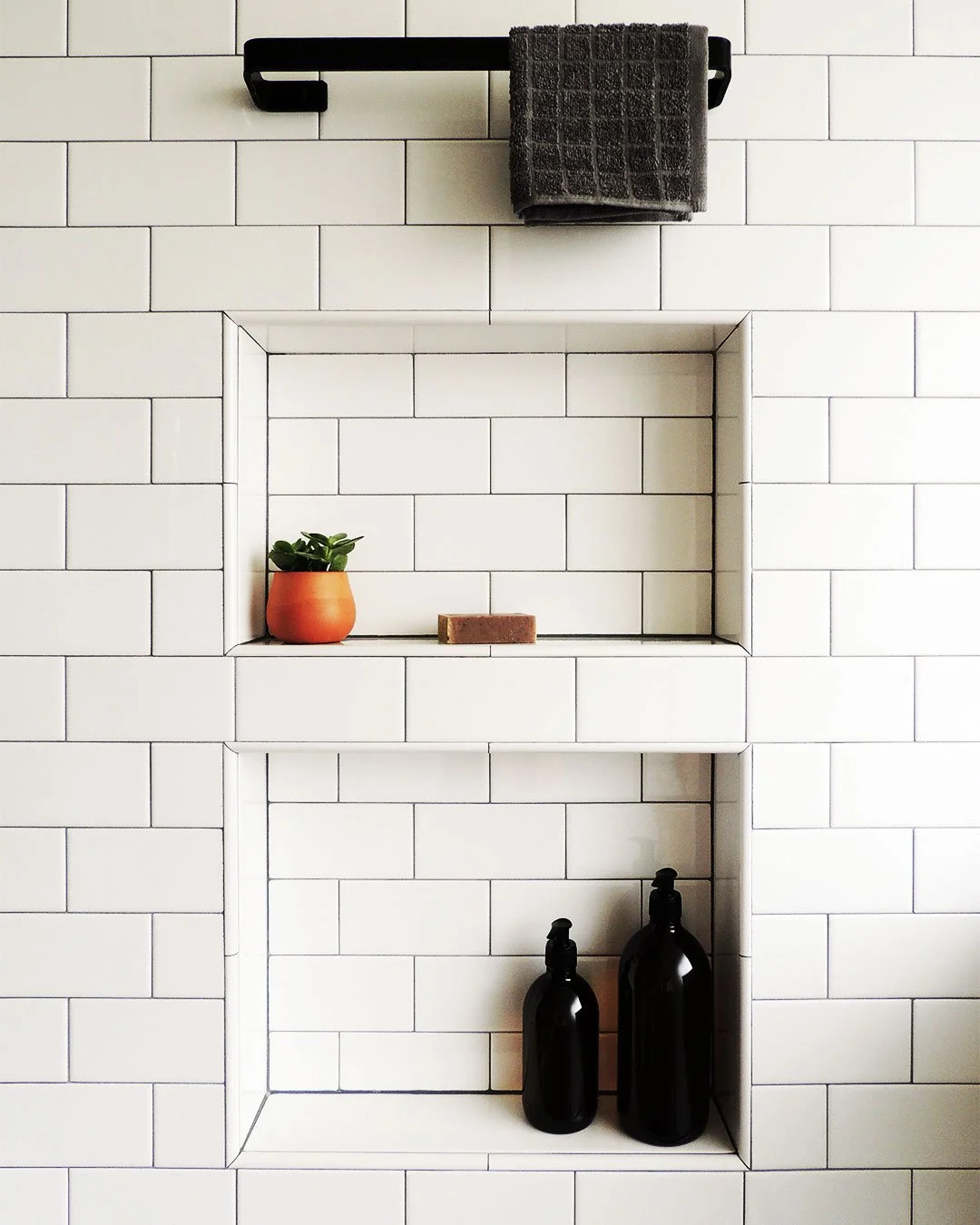 White tiled wall with built-in nook holding a potted plant and soap bottles and black metal towel bar above.