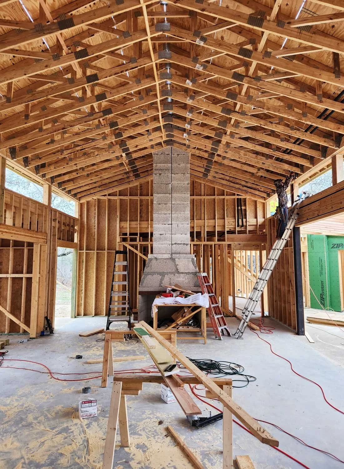 Interior of a house under construction with exposed wooden framing, a central chimney, ladders, and construction tools.