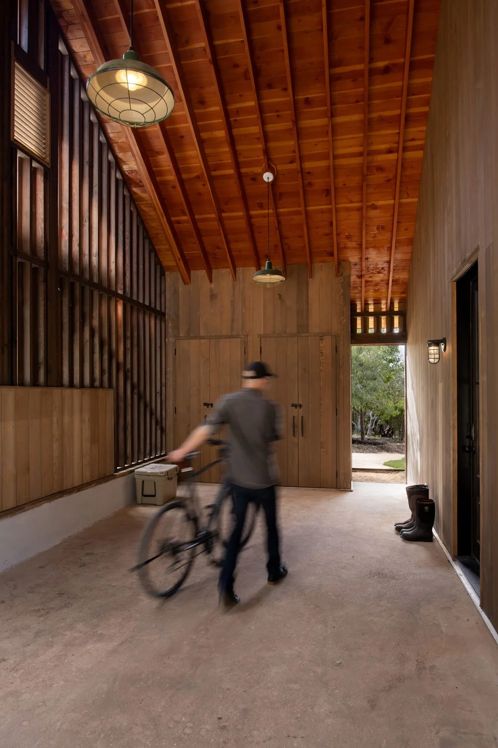 A person in motion wearing a gray shirt and black hat pushes a bicycle inside a wooden structure with a high, sloped ceiling and a doorway leading outside to a green yard.