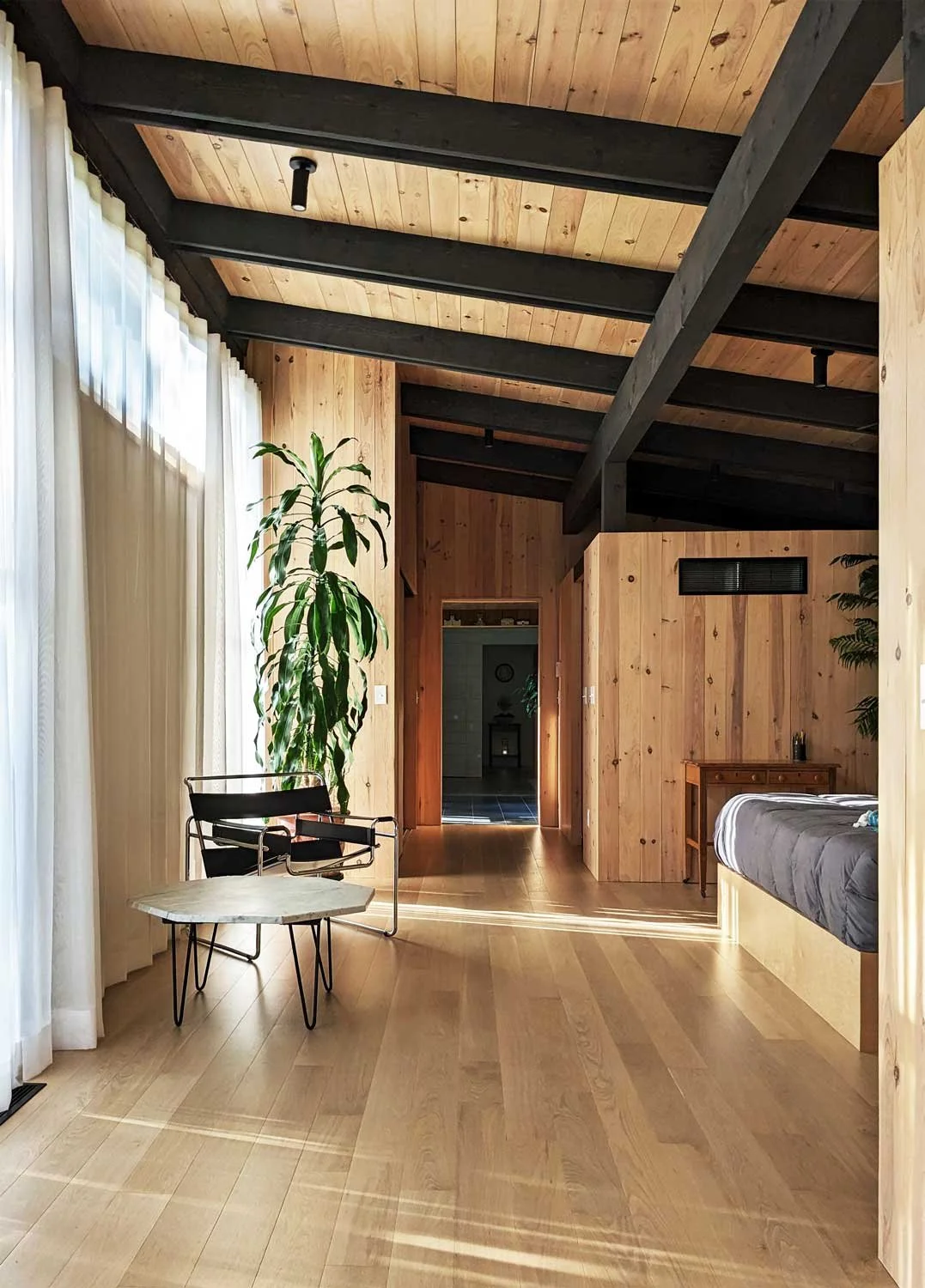 Interior view of a wooden bedroom with black ceiling beams, a large window with white curtains, and a tall green plant.