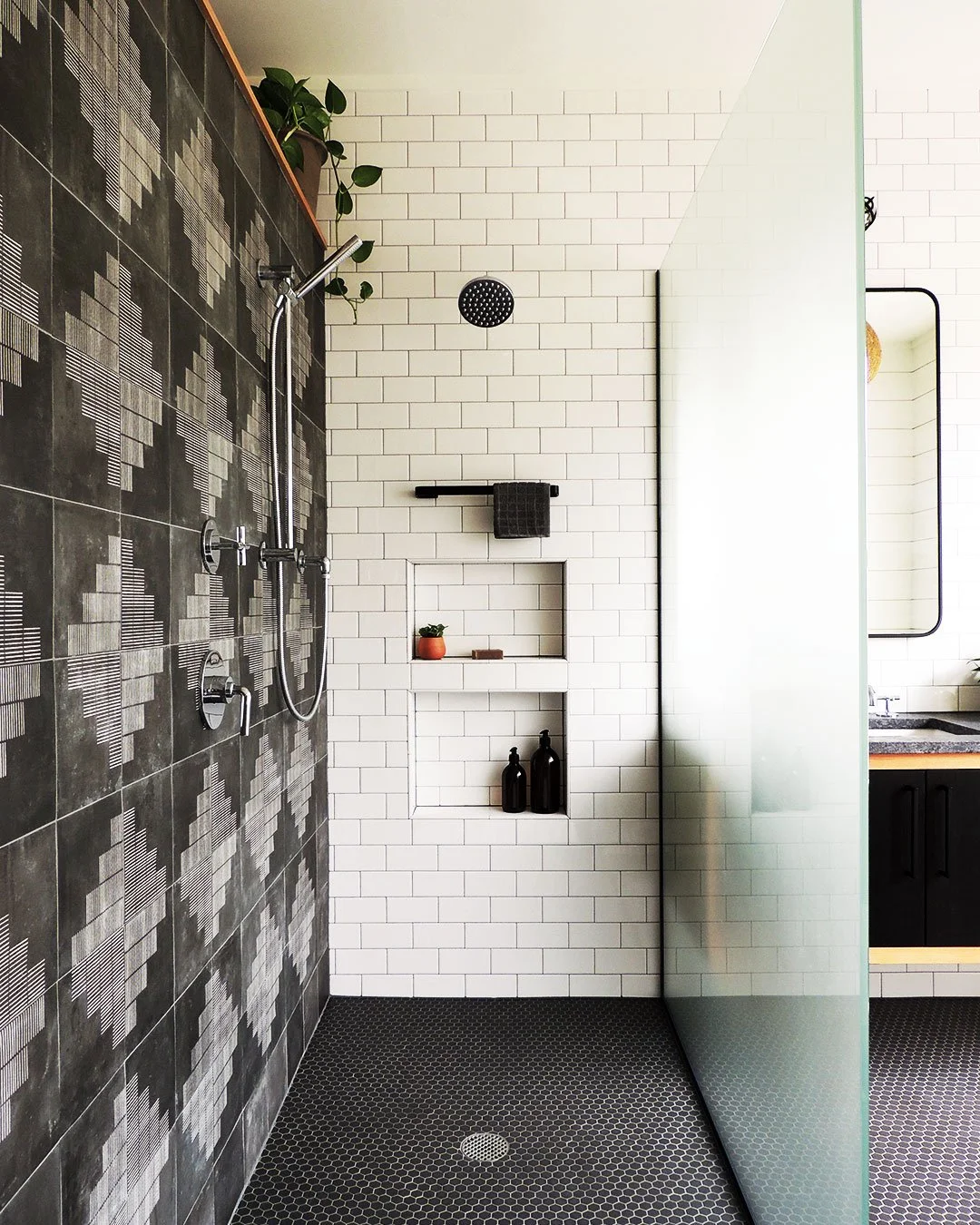 Modern bathroom with a shower area, black and gray patterned tiles on the left wall, white subway tiles on the rear wall, built-in nook with decor, and a frosted glass partition.
