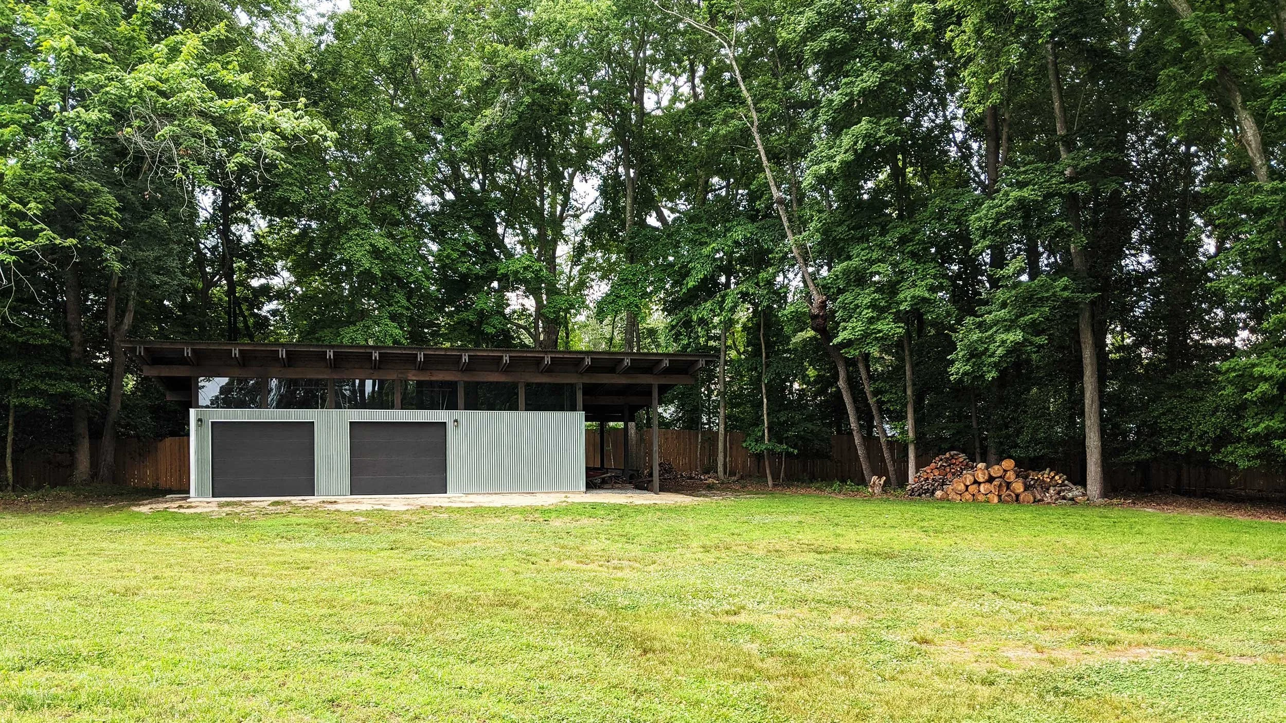 Modern shed in a grassy lawn, a woodpile, and tall trees with green foliage.