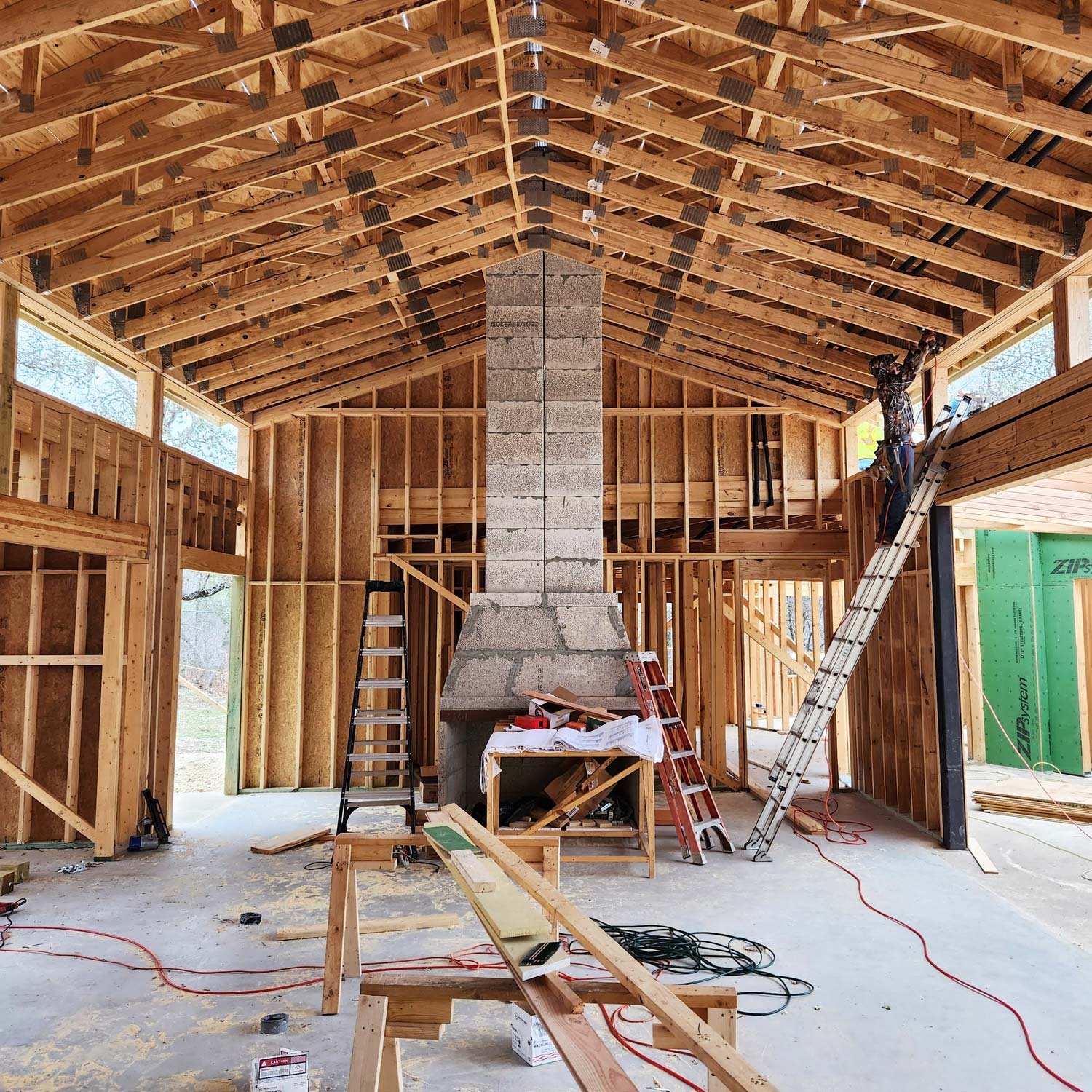 Interior of a house under construction with exposed wooden framing, a concrete chimney, and a worker on a ladder.