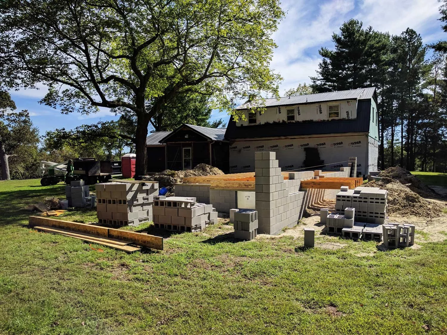 Construction site in a grassy area with stacks of concrete blocks, wooden beams, and a partially built structure near a house under renovation.