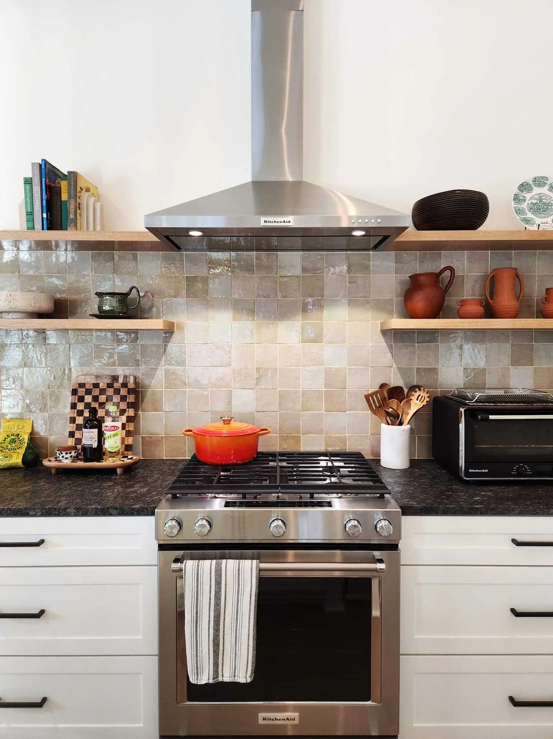 Modern kitchen with a stainless steel range and vent hood, granite countertop, floating shelves with pottery and books, and beige tile backsplash.