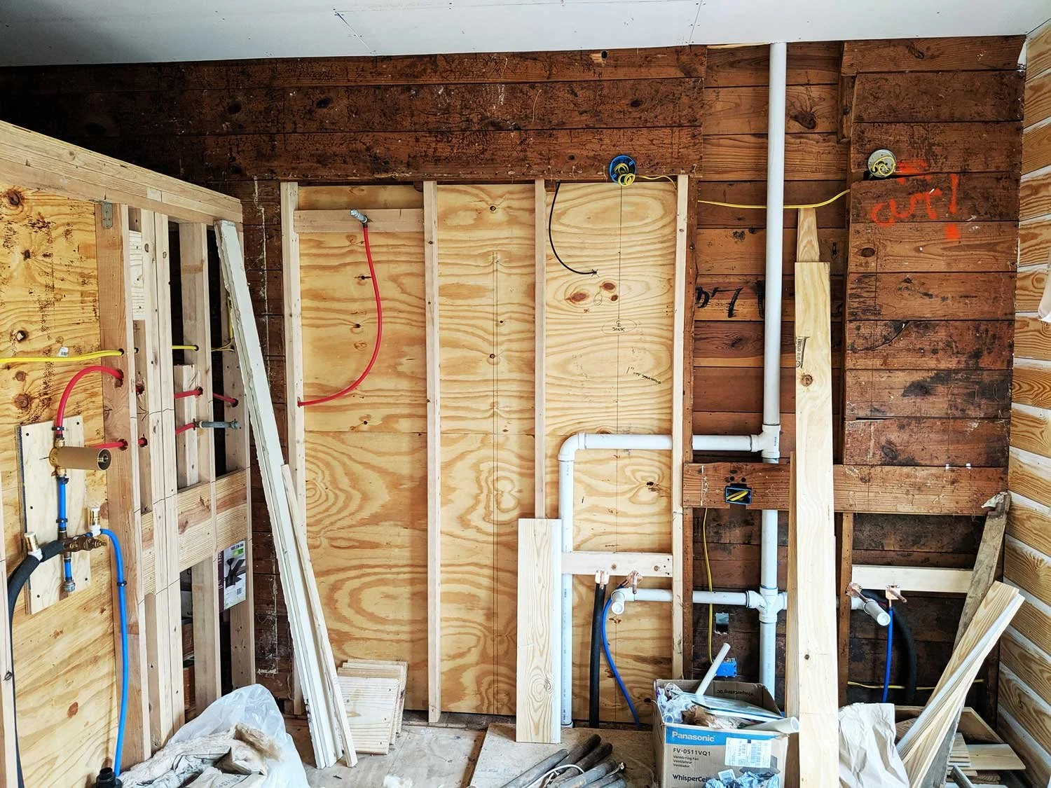 Bathroom wall under construction with plumbing pipes, wooden framing, and plywood panels.