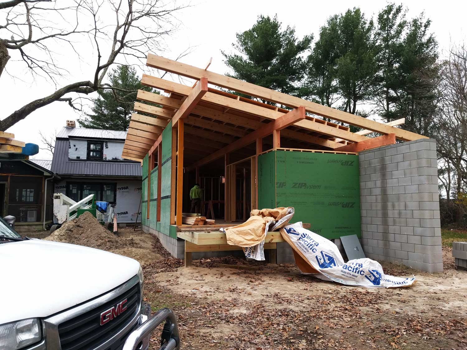 Construction site of a house with wooden framing, green sheathing, and a cinder block wall, surrounded by trees and construction materials.