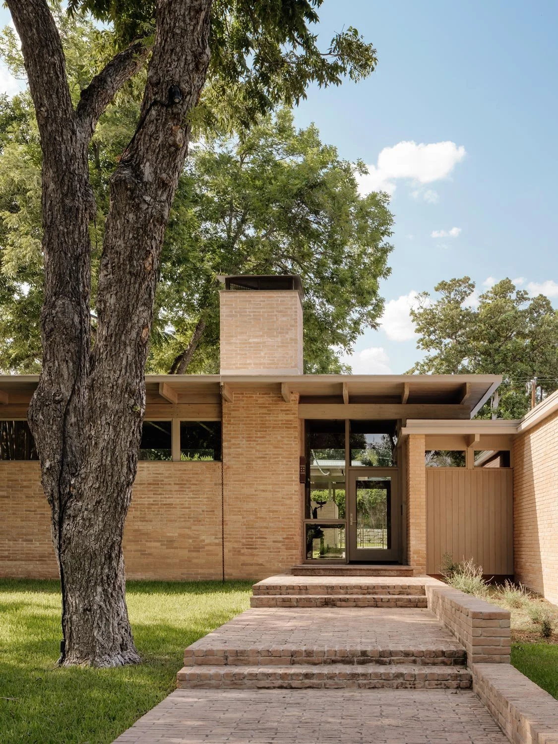 Modern brick house with large glass door, brick steps, surrounded by green lawn and tall trees.