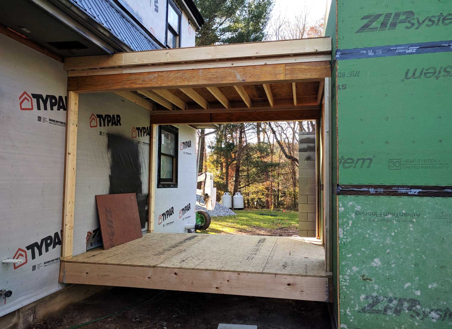 Construction site showing wooden framing and green sheathing on a building, with a view of trees and construction equipment outside.