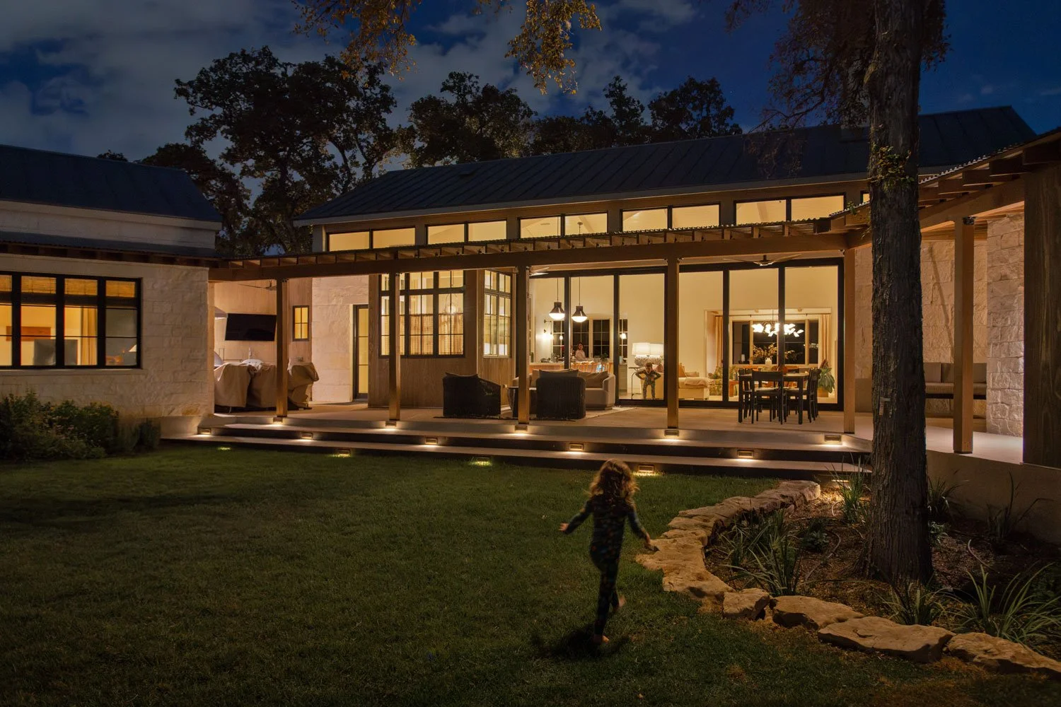Child running on a lawn in front of a house with large glass windows and a patio, illuminated at night.