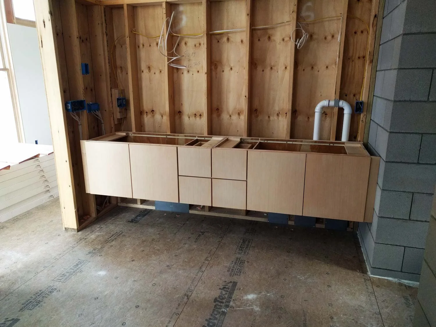 Bathroom cabinets under construction with exposed wooden framing, electrical wiring, and plumbing, and unfinished flooring.