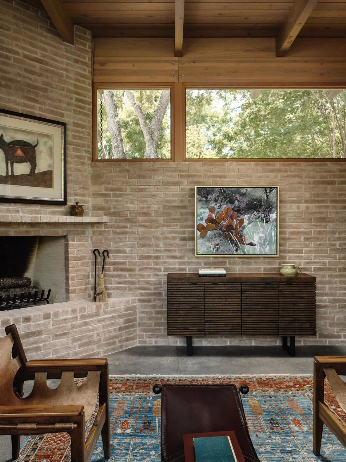 A living room with brick walls, a fireplace, a wooden ceiling, and a clerestory window showing trees outside.
